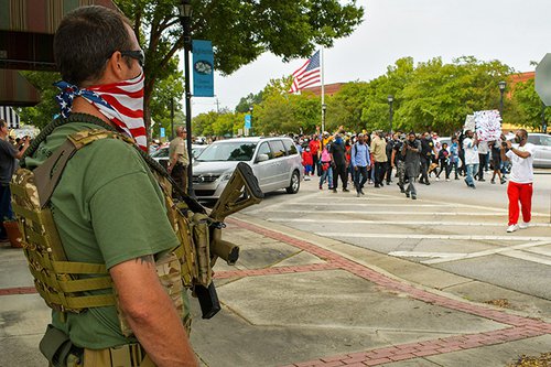 An armed bystander watches protesters march by in downtown Sylvania during the Justice Caravan for Julian Lewis on Saturday.